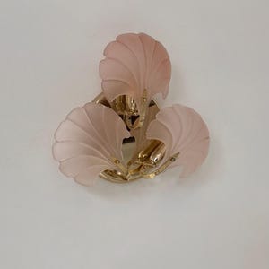 May include: A wall-mounted light fixture with three frosted pink glass shades shaped like stylized shells. The fixture has a gold-colored metal base and accents, creating a vintage aesthetic. The light is mounted on a white wall.