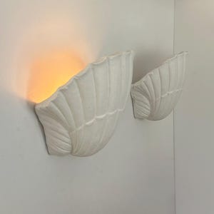 Könnte beinhalten: Zwei weiße, muschelförmige Wandleuchten. Die Leuchten sind an einer weißen Wand montiert, wobei eine beleuchtet ist und ein warmes, orangefarbenes Leuchten erzeugt. Das Design weist strukturierte, geriffelte Details auf und schafft eine dekorative Leuchte.