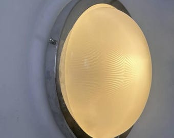 May include: A round, wall-mounted light fixture with a chrome-colored metal frame. The fixture features a frosted, semi-spherical glass shade that emits a warm, yellow light. The light is mounted on a light blue wall.