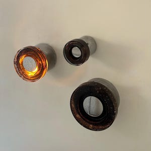 May include: Three wall-mounted light fixtures with a circular design. The fixtures have a metal base and a glass or resin lens. One fixture glows with an orange light, while the others have a dark brown lens. The fixtures are mounted on a white wall.