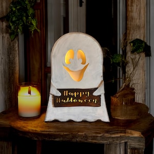 May include: A white wooden ghost-shaped Halloween decoration with a smiling face and glowing eyes and mouth. The ghost holds a sign that reads "Happy Halloween". A lit candle sits to the left of the ghost.