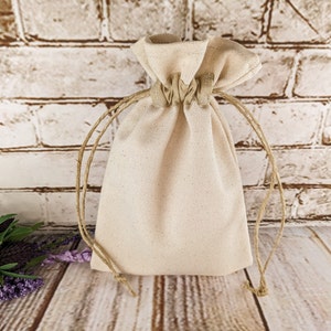 Puede incluir: Una bolsa de algod&oacute;n blanco lisa con un cord&oacute;n de yute natural. La bolsa est&aacute; vac&iacute;a y se encuentra sobre una superficie de madera marr&oacute;n claro.