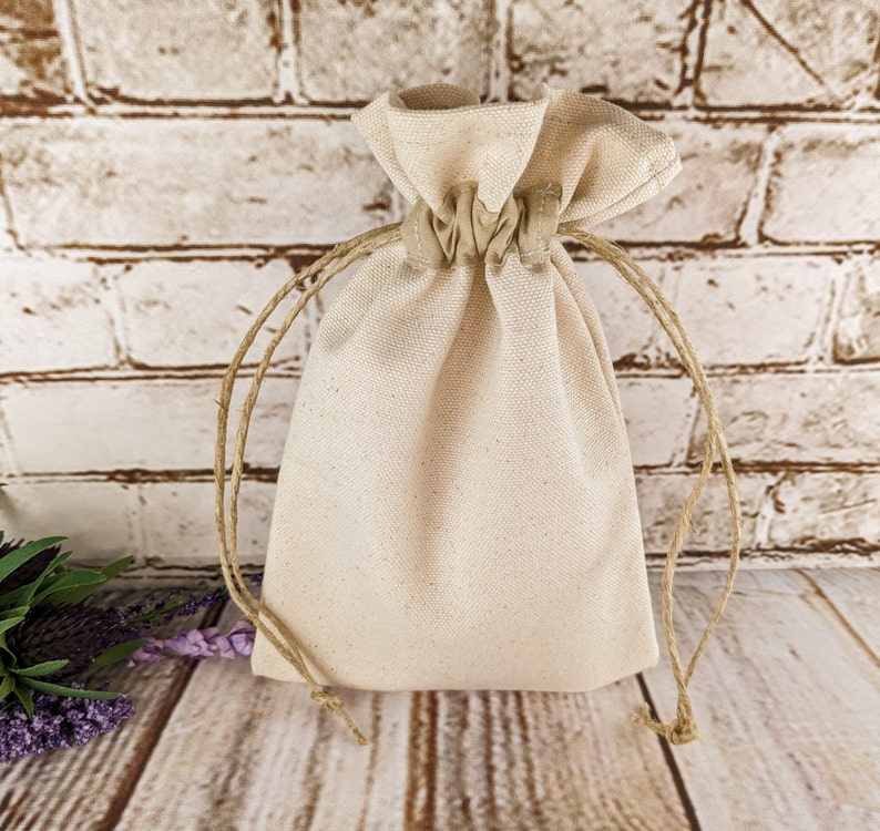 Puede incluir: Una bolsa de algod&oacute;n blanco lisa con un cord&oacute;n de yute natural. La bolsa est&aacute; vac&iacute;a y se encuentra sobre una superficie de madera marr&oacute;n claro.