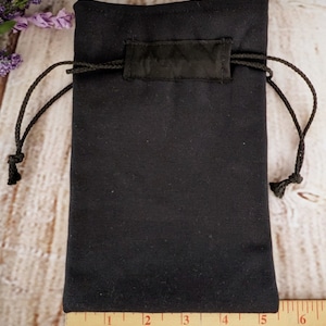 Puede incluir: Una bolsa de cord&oacute;n negro hecha de tela con una etiqueta de tela negra. La bolsa mide aproximadamente 15 cm de ancho y 20 cm de alto.