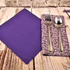 May include: A purple cloth napkin with a floral patterned napkin on top. A silver fork and spoon are placed on the floral napkin.