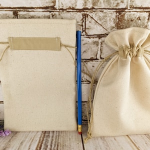 Puede incluir: Dos bolsas de algod&oacute;n de color natural con cord&oacute;n de ajuste, una con una etiqueta de tela beige, sobre una superficie de madera.