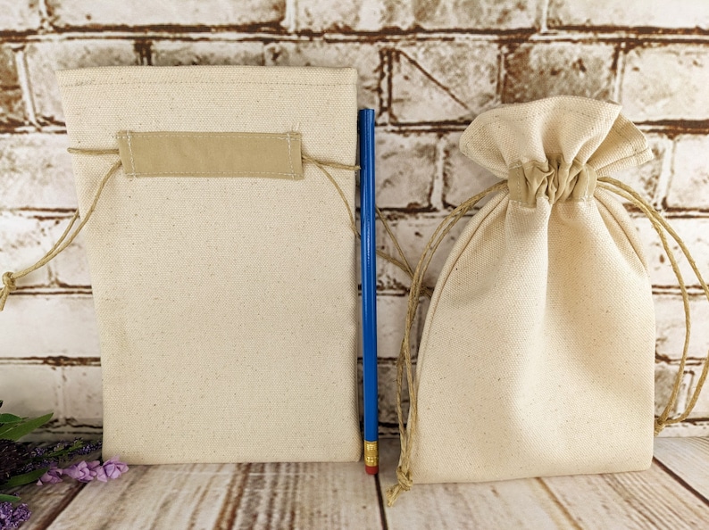 Puede incluir: Dos bolsas de algod&oacute;n de color natural con cord&oacute;n de ajuste, una con una etiqueta de tela beige, sobre una superficie de madera.