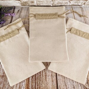 Puede incluir: Tres bolsas de algod&oacute;n de color natural con una solapa de tela marr&oacute;n y un cord&oacute;n de ajuste marr&oacute;n. Las bolsas est&aacute;n sobre una superficie de madera.