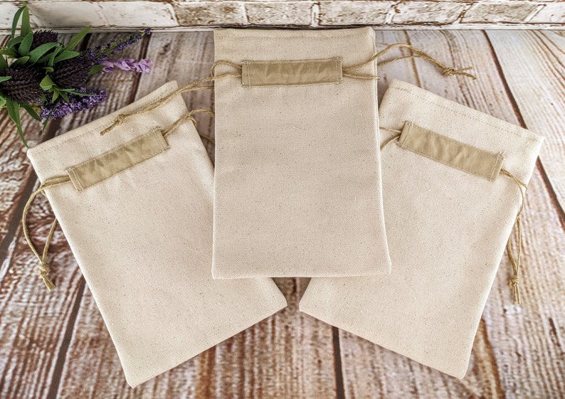 Puede incluir: Tres bolsas de algod&oacute;n de color natural con una solapa de tela marr&oacute;n y un cord&oacute;n de ajuste marr&oacute;n. Las bolsas est&aacute;n sobre una superficie de madera.