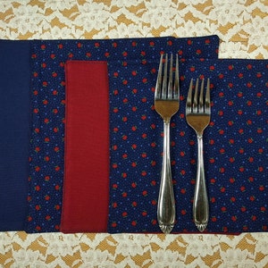 May include: Set of three fabric placemats in navy blue with a red and white apple pattern. The placemats are lined with a solid red fabric border. Two silver forks are placed on top of the placemats.