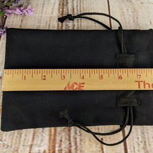 Puede incluir: Una bolsa negra de cord&oacute;n con cierre de cord&oacute;n negro. La bolsa est&aacute; tendida sobre una superficie de madera con una regla que mide 20,3 cm de largo.