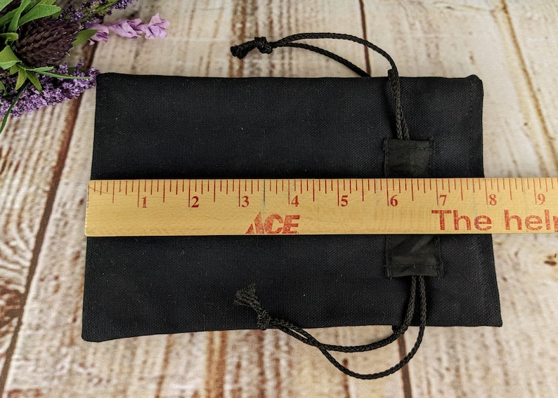 Puede incluir: Una bolsa negra de cord&oacute;n con cierre de cord&oacute;n negro. La bolsa est&aacute; tendida sobre una superficie de madera con una regla que mide 20,3 cm de largo.