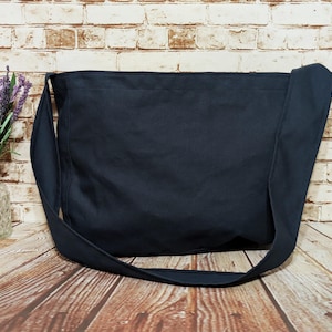 May include: A navy blue shoulder bag with a long strap, set against a brick wall and wooden surface. The bag is made of a solid, dark fabric. A vase of purple flowers is visible on the left side of the image.