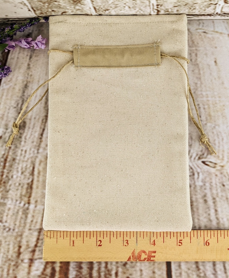 Puede incluir: Una bolsa de cord&oacute;n blanco liso con una solapa de tela beige y un cierre de cord&oacute;n. La bolsa mide 15 cm de ancho y 20 cm de alto.