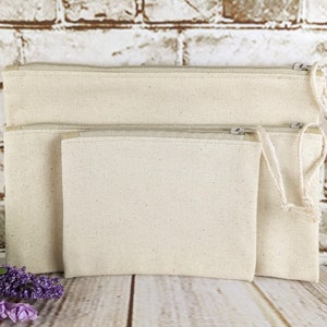 May include: Three natural canvas zippered pouches of different sizes. The pouches are made of a natural canvas fabric and have a zipper closure. The pouches are nested inside each other, with the largest pouch on the bottom and the smallest pouch on top.