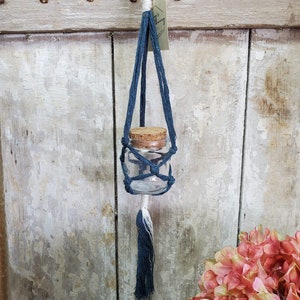 May include: A blue macrame plant hanger with a small glass jar and a cork lid. The hanger is hanging from a white wooden surface.