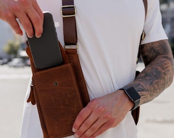 May include: A brown leather chest bag with adjustable straps and a front pocket. The bag is worn over a white t-shirt and blue jeans. A black smartphone is being placed into the bag.