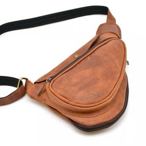 May include: A brown leather sling bag with a black adjustable strap. The bag has a curved shape with a zippered pocket on the front. The leather has a distressed look, and the bag appears to be new.