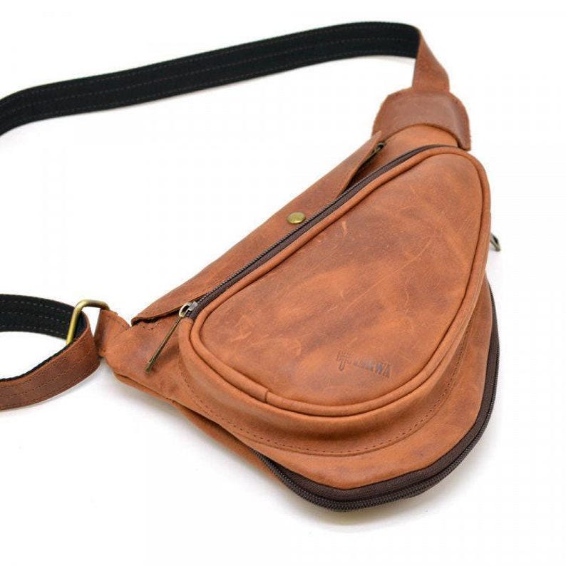 May include: A brown leather sling bag with a black adjustable strap. The bag has a curved shape with a zippered pocket on the front. The leather has a distressed look, and the bag appears to be new.