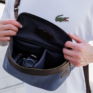 May include: A blue and brown camera bag with a shoulder strap. The bag is open, revealing a Nikon camera inside. The bag has a dark interior and brown leather trim. The person is wearing a white t-shirt.
