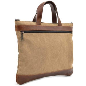 May include: A tan canvas tote bag with brown leather accents. The bag features a brown leather base, a brown leather band across the top, and brown leather handles. A black zipper runs across the top of the bag. The bag has a rectangular shape.