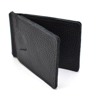 Puede incluir: Una cartera con clip para dinero de cuero negro que presenta una superficie texturizada y granulada. La cartera est&aacute; construida con un mecanismo de clip met&aacute;lico con bisagras integrado en el lomo, dise&ntilde;ado para sujetar billetes entre dos paneles.