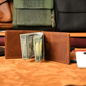 Puede incluir: Una cartera bifold de cuero marr&oacute;n con costuras visibles en los bordes, que sostiene varios billetes de cien d&oacute;lares estadounidenses en el clip central. La cartera est&aacute; abierta, mostrando su construcci&oacute;n interior y el dinero en efectivo.