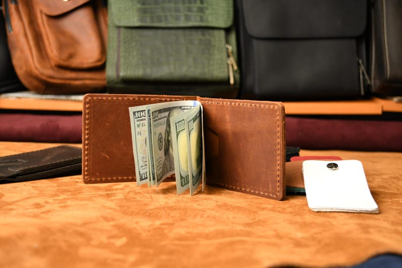 Puede incluir: Una cartera bifold de cuero marr&oacute;n con costuras visibles en los bordes, que sostiene varios billetes de cien d&oacute;lares estadounidenses en el clip central. La cartera est&aacute; abierta, mostrando su construcci&oacute;n interior y el dinero en efectivo.