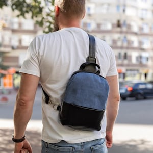 May include: A blue and black sling backpack with a single strap worn over the shoulder. The bag has a denim-like upper section and a black leather-like bottom. The strap is black and the bag is worn by a person wearing a white t-shirt and blue jeans.