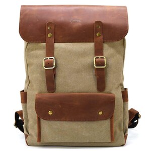 Könnte beinhalten: Ein beigefarbener Rucksack aus Segeltuch mit braunen Lederakzenten. Der Rucksack hat eine braune Lederklappe, Riemen und eine Vordertasche. Er hat goldfarbene Schnallen und Nieten. Die Tasche hat zwei Seitentaschen und verstellbare Schultergurte.