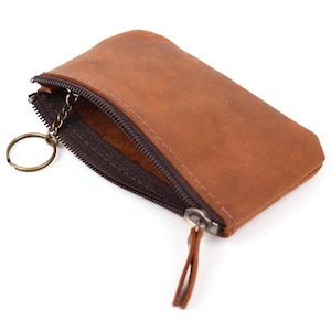 Può includere: Un portamonete in pelle color cuoio con chiusura a zip marrone scuro e tiralampo in pelle abbinato. Una catenella in metallo dorato con un anello portachiavi circolare è fissata all'interno della zip per riporre le chiavi in sicurezza.