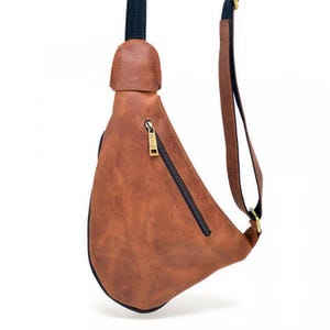 May include: A brown leather sling bag with a black zipper and strap. The bag has a textured surface and a gold-colored zipper pull. The strap is black and brown. The bag is designed to be worn across the body.