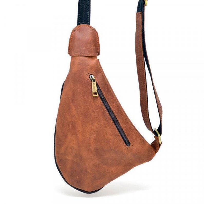May include: A brown leather sling bag with a black zipper and strap. The bag has a textured surface and a gold-colored zipper pull. The strap is black and brown. The bag is designed to be worn across the body.