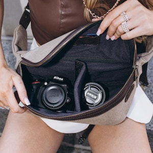 Peut inclure: Sac photo en toile beige avec garniture en cuir marron, ouvert pour révéler un appareil photo et un objectif Nikon. Le sac a une fermeture éclair et une bandoulière. L'intérieur est noir avec des séparateurs.