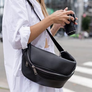 Op de afbeelding: Zwarte leren crossbodytas met een zilveren ritssluiting. De tas heeft een gebogen vorm en een lange, verstelbare riem. De tas wordt over de schouder gedragen. De achtergrond is een stadsstraat.