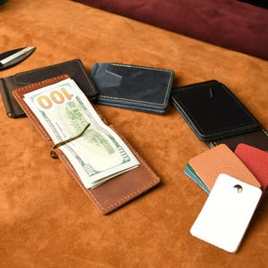 Puede incluir: Una colecci&oacute;n de carteras con clip para billetes de cuero en varios colores, incluyendo marr&oacute;n, negro y azul, con una cartera marr&oacute;n que contiene un billete de cien d&oacute;lares. Se incluyen unas tijeras de metal negro y muestras de cuero texturizado.