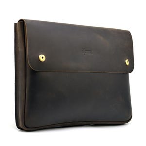 May include: A dark brown leather laptop sleeve with a flap closure secured by two brass-colored snap closures. The sleeve has a rectangular shape and a subtle embossed logo near the top. The leather has a distressed appearance.