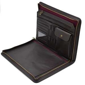 May include: Open brown leather portfolio with multiple interior pockets, including card slots, pen holders, and a small pouch with a button closure. The portfolio has a full-length zipper with a gold-colored pull and a burgundy interior lining.