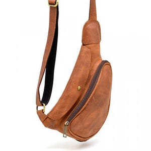 May include: A brown leather sling bag with a curved design. The bag features a zippered main compartment, a small front pocket, and an adjustable shoulder strap. The bag has brass-colored hardware and a textured finish.