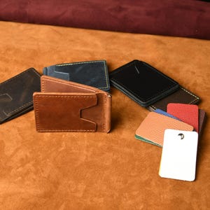 Puede incluir: Una colecci&oacute;n de carteras con clip para billetes de cuero en marr&oacute;n, negro y azul marino, con costuras visibles y ranuras para tarjetas. Junto a las carteras hay varias muestras de cuero rectangulares peque&ntilde;as de colores y una etiqueta blanca.
