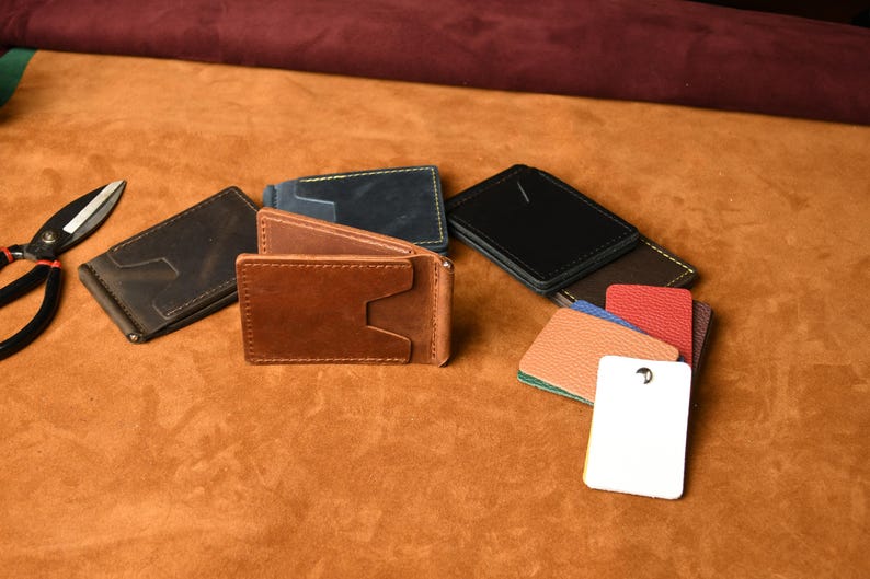 Puede incluir: Una colecci&oacute;n de carteras con clip para billetes de cuero en marr&oacute;n, negro y azul marino, con costuras visibles y ranuras para tarjetas. Junto a las carteras hay varias muestras de cuero rectangulares peque&ntilde;as de colores y una etiqueta blanca.