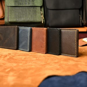 Puede incluir: Una fila de cinco carteras rectangulares de cuero con clip para billetes en varios colores, incluyendo marr&oacute;n oscuro, azul, tostado, negro y marr&oacute;n medio. Cada cartera presenta costuras visibles en los bordes y un clip met&aacute;lico en el lateral.