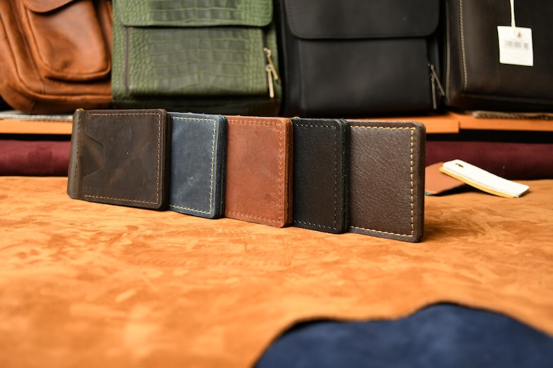 Puede incluir: Una fila de cinco carteras rectangulares de cuero con clip para billetes en varios colores, incluyendo marr&oacute;n oscuro, azul, tostado, negro y marr&oacute;n medio. Cada cartera presenta costuras visibles en los bordes y un clip met&aacute;lico en el lateral.