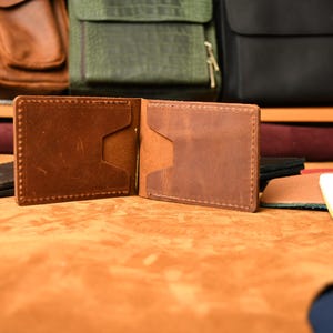 Puede incluir: Una cartera de cuero marr&oacute;n tipo bifold con un clip met&aacute;lico central para billetes, costuras visibles en los bordes y dos ranuras para tarjetas en cada lado interior. La cartera est&aacute; hecha de cuero liso con acabado mate y textura natural.