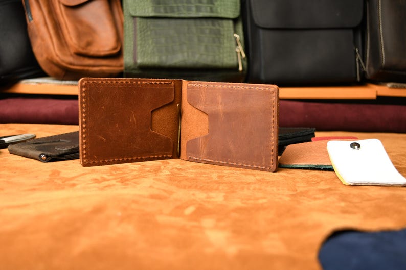 Puede incluir: Una cartera de cuero marr&oacute;n tipo bifold con un clip met&aacute;lico central para billetes, costuras visibles en los bordes y dos ranuras para tarjetas en cada lado interior. La cartera est&aacute; hecha de cuero liso con acabado mate y textura natural.