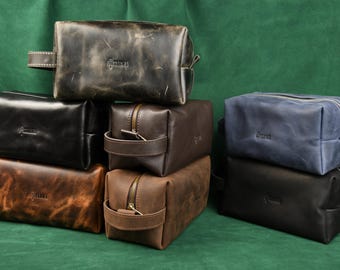 May include: A collection of rectangular leather toiletry bags in various colors including black, brown, and blue. Each bag features a side handle, a top zipper closure, and the embossed Tarwa logo on the front surface of the leather material.