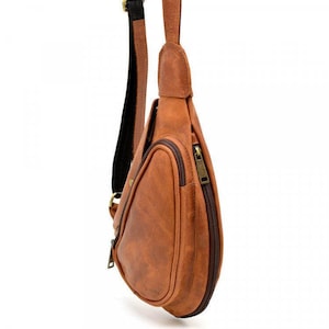 May include: A brown leather sling bag with a zippered main compartment and an exterior pocket. The bag has a black and brown adjustable strap and a bronze-colored zipper. The bag is designed to be worn across the body.