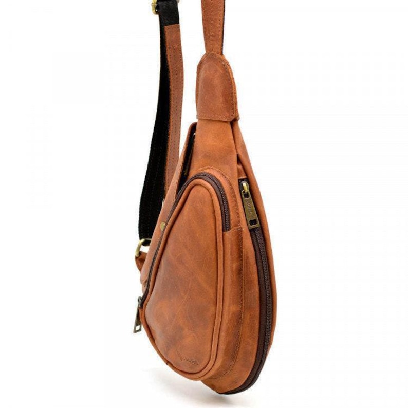 May include: A brown leather sling bag with a zippered main compartment and an exterior pocket. The bag has a black and brown adjustable strap and a bronze-colored zipper. The bag is designed to be worn across the body.