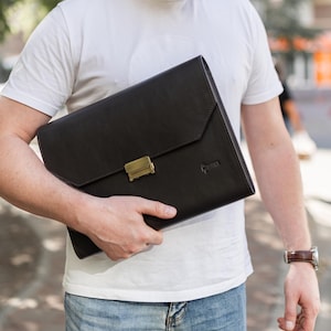 May include: A dark brown leather portfolio with a gold-colored clasp. The portfolio is rectangular with a flap closure. The word "PARKER" is embossed on the lower right corner. The portfolio is being held by a person wearing a white t-shirt and blue jeans.