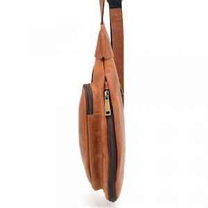 May include: A brown leather crossbody bag with a zippered main compartment and a smaller front pocket. The bag has a brown strap and a gold-colored zipper. The bag is hanging against a white background.